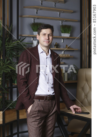 Young man in a burgundy suit with a mobile phone standing in the office and looks at the camera. Dark hair, brown eyes, white skin, a smile. In the background there are shelves with books and flowers. Young man in a burgundy suit with a mobile phone standing in the office and looks at the camera. Dark hair, brown eyes, white skin, a smile. In the background there are shelves with books and flowers. 85167903
