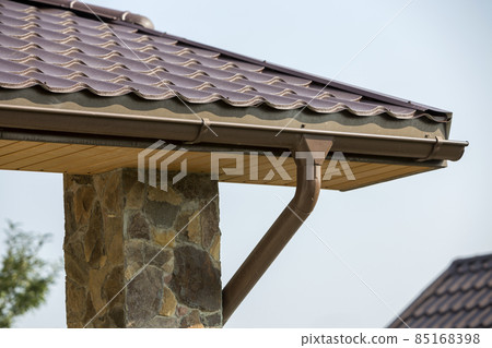 Detail of new modern house cottage corner with stucco walls decorated with natural stones, brown shingled roof and rain gutter pipe system on blue sky copy space background. Real estate concept. 85168398
