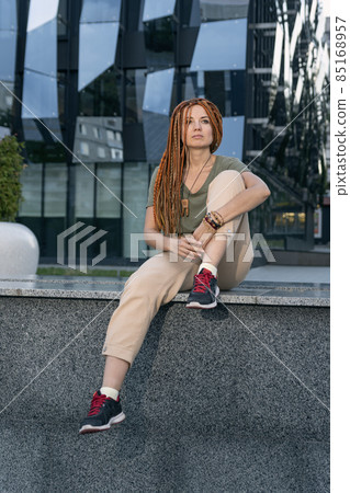 Girl with dreadlocks sits on parapet and looks thoughtfully into the distance.Woman next to building with unusual design Girl with dreadlocks sits on parapet and looks thoughtfully into the distance.Woman next to building with unusual design 85168957