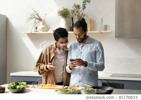 Happy young African American couple using cellphones, cooking in kitchen. Happy young African American couple using cellphones, cooking in kitchen. 85170633