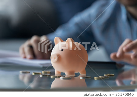Man calculates expenses, piggybank and coins on foreground Man calculates expenses, piggybank and coins on foreground 85170840
