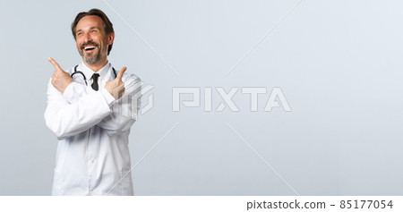 Covid-19, coronavirus outbreak, healthcare workers and pandemic concept. Carefree cheerful male doctor laughing over promo at top left. Therapist in white coat pointing sideways at advertisement 85177054