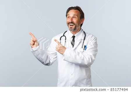 Covid-19, coronavirus outbreak, healthcare workers and pandemic concept. Happy smiling male doctor in white coat pointing fingers upper left corner amused, showing medication or clinic banner 85177494