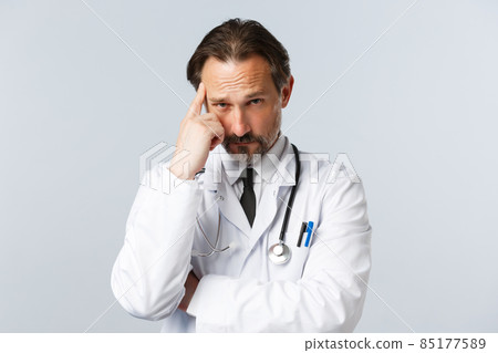 Covid-19, preventing virus, healthcare workers and vaccination concept. Thoughtful serious and skeptical doctor in white coat, pointing at temple, worried about patient health, white background 85177589