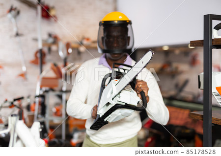 African-american man in protective helmet and with chainsaw in hands in gardening tools shop. 85178528