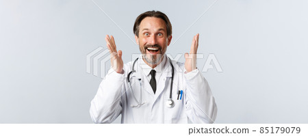 Covid-19, preventing virus, healthcare workers and vaccination concept. Close-up of excited bearded male doctor in white coat raising hands up thrilled, hear great news and rejoicing 85179079