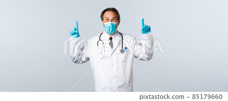 Covid-19, preventing virus, healthcare workers and vaccination concept. Impressed surprised male doctor in medical mask and gloves, pointing fingers up to show great news Covid-19, preventing virus, healthcare workers and vaccination concept. Impressed surprised male doctor in medical mask and gloves, pointing fingers up to show great news 85179660