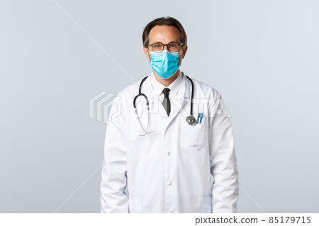 Covid-19, preventing virus, healthcare workers and vaccination concept. Serious confident male doctor in white coat and medical mask, wearing glasses, ready treat patient with influenza Covid-19, preventing virus, healthcare workers and vaccination concept. Serious confident male doctor in white coat and medical mask, wearing glasses, ready treat patient with influenza 85179715