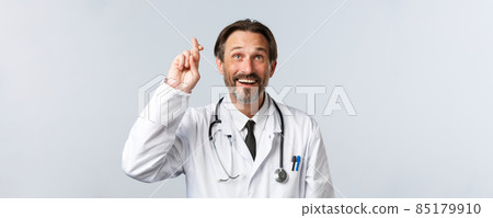 Covid-19, preventing virus, healthcare workers and vaccination concept. Close-up of optimistic hopeful doctor in white coat, cross fingers good luck and looking up smiling, making wish Covid-19, preventing virus, healthcare workers and vaccination concept. Close-up of optimistic hopeful doctor in white coat, cross fingers good luck and looking up smiling, making wish 85179910