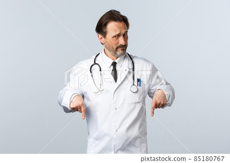 Covid-19, coronavirus outbreak, healthcare workers and pandemic concept. Skeptical and displeased male doctor in white coat, looking doubtful pointing fingers down 85180767
