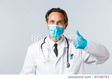 Covid-19, preventing virus, healthcare workers and vaccination concept. Excited happy doctor smiling enthusiastic, wear medical mask and gloves show thumbs-up, agree or approve product 85180791