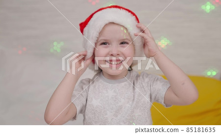 little child laughs in Santa hat, kid smile Christmas holidays, happy family, baby has fun playing bright colors of garland, festive mood in new year, waiting for miracle child dream, time for gifts 85181635