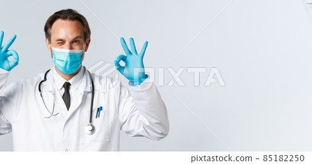 Covid-19, preventing virus, healthcare workers and vaccination concept. Friendly smiling doctor in medical mask and gloves, wink cheeky and show okay gesture, guarantee or recommend service 85182250