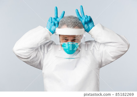 Covid-19, preventing virus, healthcare workers and vaccination concept. Happy optimistic doctor in personal protective equipment showing bunny ears gesture with fingers up above head 85182269