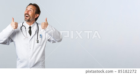 Covid-19, coronavirus outbreak, healthcare workers and pandemic concept. Enthusiastic handsome doctor in white coat looking at top left advertisement and thumbs-up, recommend product Covid-19, coronavirus outbreak, healthcare workers and pandemic concept. Enthusiastic handsome doctor in white coat looking at top left advertisement and thumbs-up, recommend product 85182272