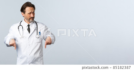 Covid-19, coronavirus outbreak, healthcare workers and pandemic concept. Skeptical and displeased male doctor in white coat, looking doubtful pointing fingers down 85182387
