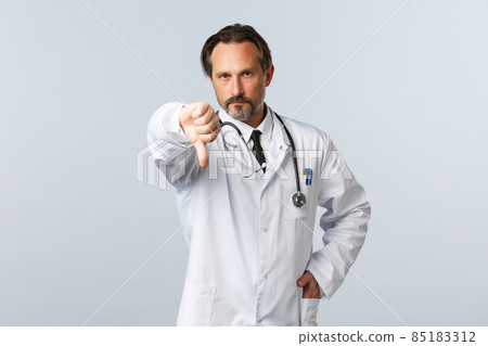 Covid-19, coronavirus outbreak, healthcare workers and pandemic concept. Disappointed serious male doctor in white coat, frowning displeased and show thumb-down, disapprove action Covid-19, coronavirus outbreak, healthcare workers and pandemic concept. Disappointed serious male doctor in white coat, frowning displeased and show thumb-down, disapprove action 85183312