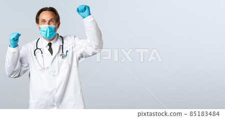 Covid-19, preventing virus, healthcare workers and vaccination concept. Relieved supportive doctor in medical mask and gloves, raising hands up as chanting, celebrating achievement of goal 85183484