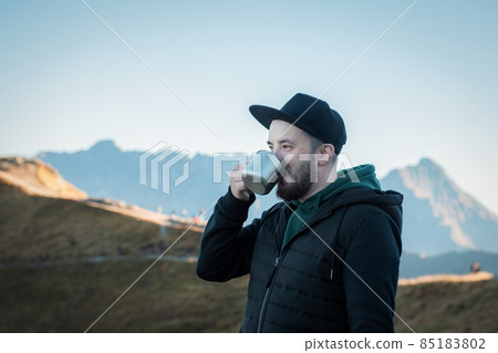 Bearded traveler man drinks tea or coffee from  metal cup while resting on a rock 85183802