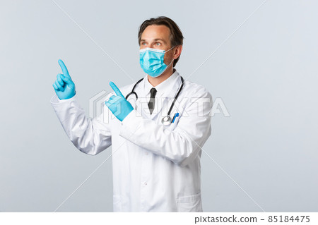 Covid-19, preventing virus, healthcare workers and vaccination concept. Middle-aged male doctor in white coat, medical mask and gloves looking upper left corner at pointing banner 85184475