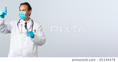 Covid-19, preventing virus, healthcare workers and vaccination concept. Serious professional med lab doctor in medical mask and gloves looking at test-tube with coronavirus vaccine 85184478