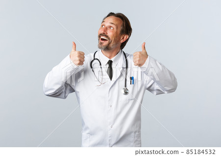 Covid-19, coronavirus outbreak, healthcare workers and pandemic concept. Enthusiastic handsome doctor in white coat looking at top left advertisement and thumbs-up, recommend product 85184532