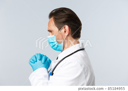 Covid-19, preventing virus, healthcare workers and vaccination concept. Close-up profile of doctor in medical mask and gloves, clench hands in plead, praying for patients health, supplicating 85184955