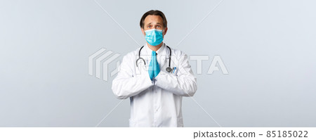 Covid-19, preventing virus, healthcare workers and vaccination concept. Hopeful worried doctor in medical mask and gloves holding hands in pray, frowning as begging, pleading favour Covid-19, preventing virus, healthcare workers and vaccination concept. Hopeful worried doctor in medical mask and gloves holding hands in pray, frowning as begging, pleading favour 85185022