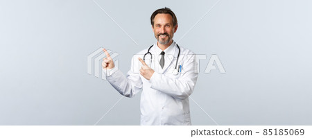 Covid-19, coronavirus outbreak, healthcare workers and pandemic concept. Smiling friendly-looking doctor in white coat pointing fingers upper left corner, showing advertisement Covid-19, coronavirus outbreak, healthcare workers and pandemic concept. Smiling friendly-looking doctor in white coat pointing fingers upper left corner, showing advertisement 85185069