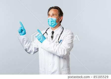 Covid-19, preventing virus, healthcare workers and vaccination concept. Upset and worried male doctor in medical mask and gloves looking anxious, panic pointing upper left corner, stare at banner Covid-19, preventing virus, healthcare workers and vaccination concept. Upset and worried male doctor in medical mask and gloves looking anxious, panic pointing upper left corner, stare at banner 85185529
