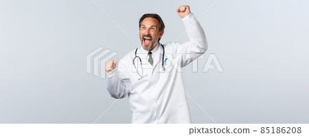 Covid-19, coronavirus outbreak, healthcare workers and pandemic concept. Enthusiastic male doctor in white coat raising hand up and shouting yes, watching sport match, winning, triumphing 85186208