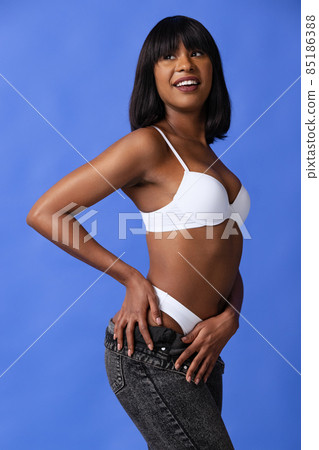Emotional adorable dark skinned girl in white underwear and jeans posing isolated over blue background. Natural beauty concept. 85186388