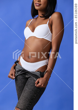 Cropped image of emotional adorable dark skinned girl in white underwear and jeans posing isolated over blue background. 85186389