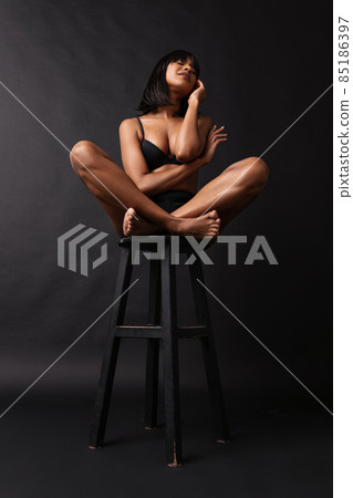 Portrait of young adorable dark skinned girl in black underwear sitting on high chair isolated over dark background. Natural beauty concept. Portrait of young adorable dark skinned girl in black underwear sitting on high chair isolated over dark background. Natural beauty concept. 85186397