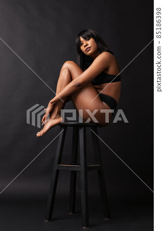 Portrait of young adorable dark skinned girl in black underwear sitting on high chair isolated over dark background. Natural beauty concept. Portrait of young adorable dark skinned girl in black underwear sitting on high chair isolated over dark background. Natural beauty concept. 85186398
