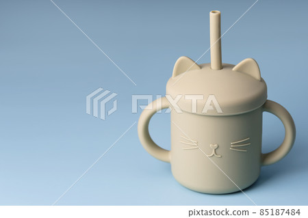 Green sippy cup on blue background. Silicone baby dishes, first soft cup for toddler. Baby tableware. Nutrition and feeding concept. Free space for text Green sippy cup on blue background. Silicone baby dishes, first soft cup for toddler. Baby tableware. Nutrition and feeding concept. Free space for text 85187484