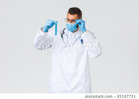 Covid-19, healthcare workers, pandemic and preventing virus concept. Thoughtful and intrigued doctor in medical mask and gloves, look interested at test-tube with new coronavirus vaccine 85187731