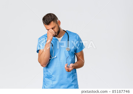 Healthcare workers, medicine, covid-19 and pandemic self-quarantine concept. Tired young doctor, male nurse during night shift in clinic helping patients with coronavirus, rub eyes exhausted 85187746
