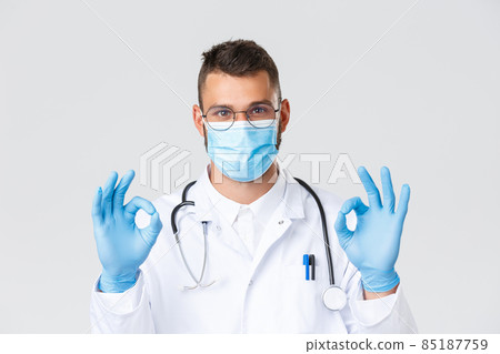 Covid-19, healthcare workers, pandemic and preventing virus concept. Close-up of satisfied handsome doctor in white coat and glasses, physician in medicla mask show okay sign, recommend promo 85187759