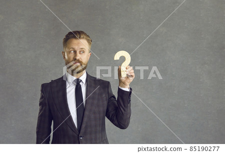 Confused puzzled businessman holding wooden question mark standing on gray on background. Pensive man in a suit is looking for answers to questions when planning a new business development strategy. 85190277