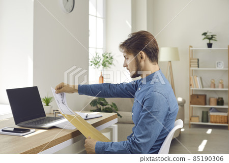 Young man who's sitting at desk in his home office opens envelope with business letter. Serious businessman sitting at table with laptop computer takes and reads sales report that he got by paper mail Young man who's sitting at desk in his home office opens envelope with business letter. Serious businessman sitting at table with laptop computer takes and reads sales report that he got by paper mail 85190306