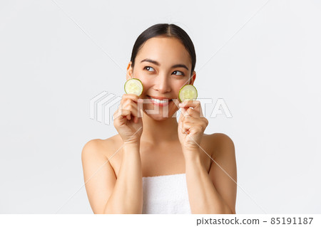 Beauty, personal care, spa salon and skincare concept. Coquettish beautiful asian girl in bath towel looking away dreamy with smile, showing cucumber pieces as promote face cream or mask Beauty, personal care, spa salon and skincare concept. Coquettish beautiful asian girl in bath towel looking away dreamy with smile, showing cucumber pieces as promote face cream or mask 85191187