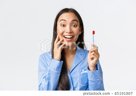 Excited and happy gorgeous asian girl in blue pajama looking amazed and satisfied with white teeth smiling at camera while holding toothbrush, found perfect toothpaste, white background Excited and happy gorgeous asian girl in blue pajama looking amazed and satisfied with white teeth smiling at camera while holding toothbrush, found perfect toothpaste, white background 85191807