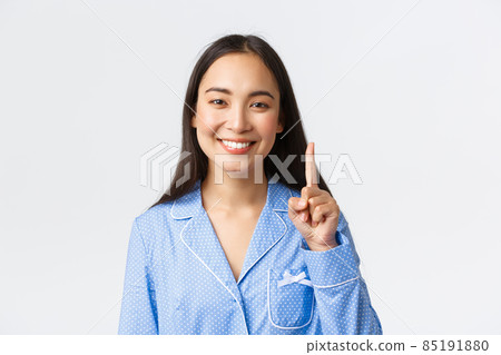 Close-up shot of happy attractive asian woman in blue pyjama showing number one, one finger and smiling white teeth, explain main rule or concept, standing white background 85191880