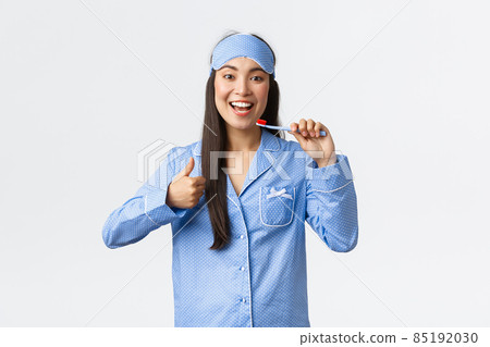 Hygiene, lifestyle and people at home concept. Pleased happy asian girl in jammies and sleeping mask, showing thumbs up while brushing white teeth with toothbrush, recommend toothpaste 85192030