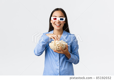 Home leisure, sleepover and slumber party concept. Smiling pleased asian girl enjoying movie night in her pajamas, wearing 3d glasses, eating popcorn, standing white background 85192032