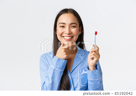 Close-up of attractive asian woman in blue pajama thinking while brushing her teeth with toothbrush, smiling and touching chin pondering great idea, white background Close-up of attractive asian woman in blue pajama thinking while brushing her teeth with toothbrush, smiling and touching chin pondering great idea, white background 85192042