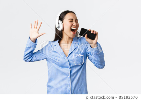 Home leisure, weekends and lifestyle concept. Excited and carefree asian girl in pajama, playing karaoke app on smartphone, singing song into mobile phone as wearing headphones, white background Home leisure, weekends and lifestyle concept. Excited and carefree asian girl in pajama, playing karaoke app on smartphone, singing song into mobile phone as wearing headphones, white background 85192570