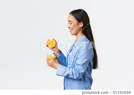 Morning, active and healthy lifestyle and home concept. Profile or beautiful healthy asian girl in blue pajamas squeezing orange juice in glass and smiling happy, starting day right Morning, active and healthy lifestyle and home concept. Profile or beautiful healthy asian girl in blue pajamas squeezing orange juice in glass and smiling happy, starting day right 85192699