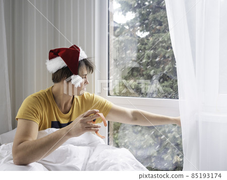 Man in Santa hat is waking up after Christmas party. He is suffering of headache after New Year celebration. Early heavy morning. Alcohol withdrawal. Worst hangover. Snowfall outdoors. 85193174